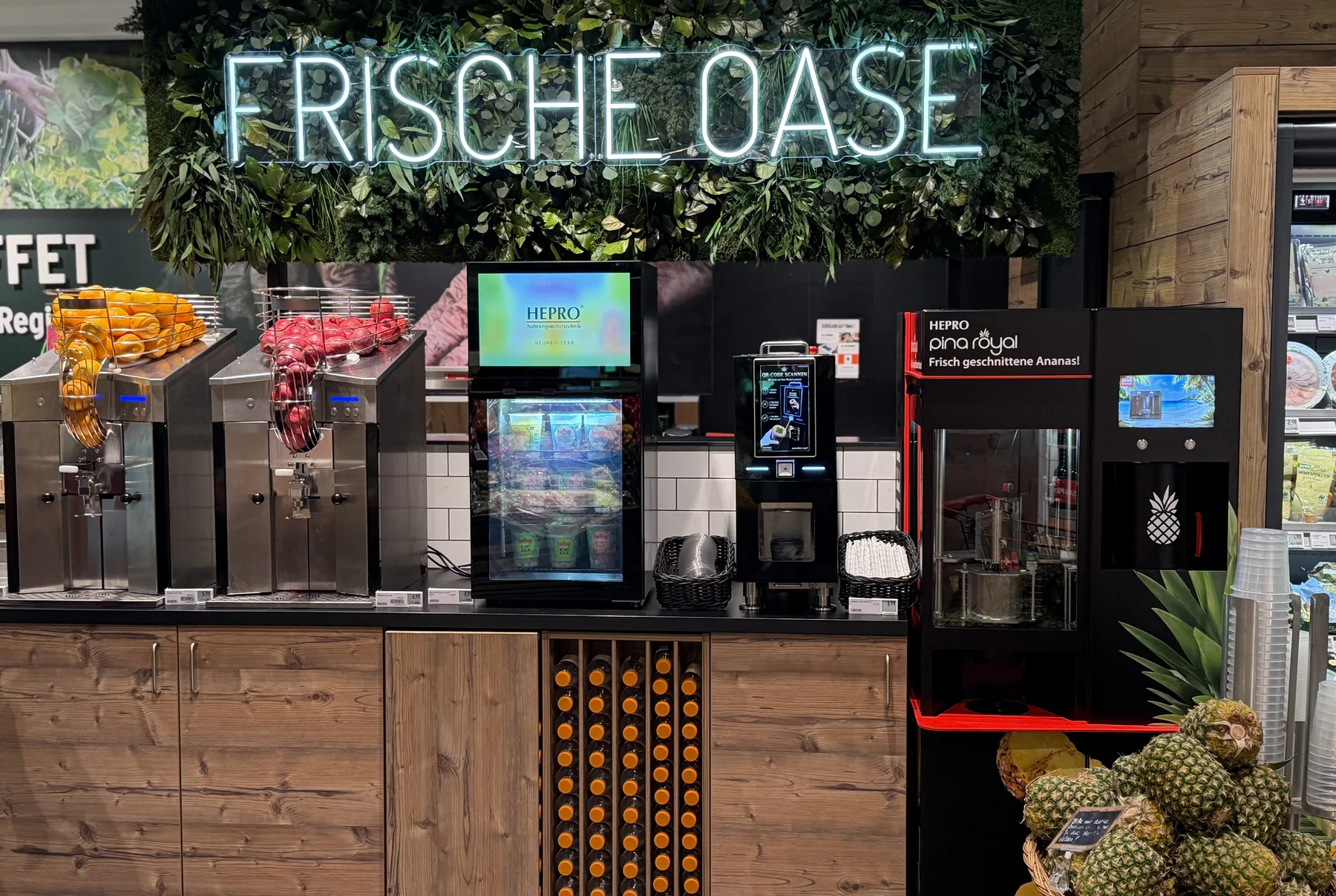 Theke „Frische Oase“ im Markt: Saftpressen, Smoothie-Automat und pina royal unter einer Leuchtschrift in einer begrünten Wand